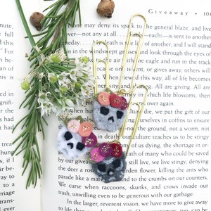 May include: Three felt skull-shaped embellishments with floral accents and gold paperclip attachments. The skulls are gray, white, and dark gray, with pink, red, and gold flower details. The background includes a book and greenery.
