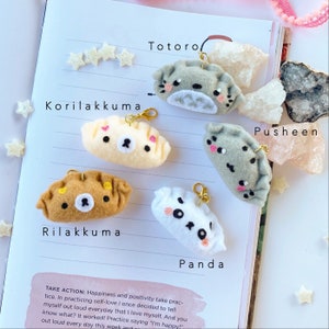 May include: Five felt plushie keychains shaped like dumplings, each featuring a different character from popular Japanese animation. The characters are Totoro, Korilakkuma, Rilakkuma, Pusheen, and Panda.