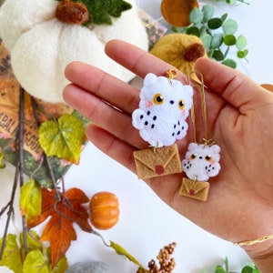 May include: Two white felt owl charms with brown felt envelopes attached. The owls have black eyes and pink cheeks. The charms are attached to gold paper clips.