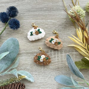 Op de afbeelding: Drie vilten wolk-vormige bedels in wit, bruin en beige, elk versierd met bloemenborduurwerk en een goudkleurige sluiting. De bedels zijn gerangschikt op een houten oppervlak, omringd door decoratief gebladerte en een dennenappel.