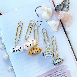 May include: Five gold paper clips with felt animal charms attached. The charms are a white dog, a beige dog, a brown dog, a gray cat, and a gray cat. The charms are all in the shape of a dumpling.