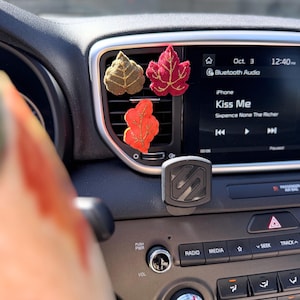 May include: Three felt leaf car air fresheners in shades of brown, red, and orange are clipped to a car vent. The car's dashboard displays the time, date, and the song "Kiss Me" by Sixpence None The Richer.