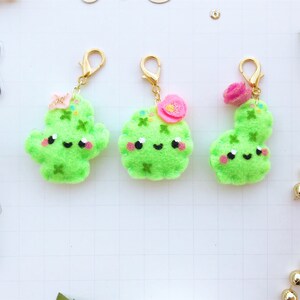 May include: Three green felt cactus keychains with gold key rings. Each cactus has a happy face and pink flower embellishments.