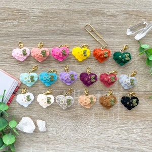 May include: A collection of 18 felt heart charms with gold accents and floral designs in various colors, including pink, purple, blue, green, red, white, and yellow. The charms are attached to gold jump rings and are perfect for adding a touch of whimsy to your crafts.