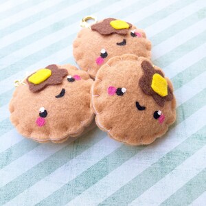 May include: Three felt pancake keychains with smiling faces, brown pancake base, brown and yellow butter, and pink cheeks. Each keychain has a gold clasp. The keychains are on a light green and white striped background.
