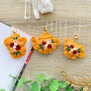 Peut inclure: Trois breloques en feutre en forme de théière orange, ornées de broderies florales rouges, roses et blanches. Chaque breloque est dotée d'un fermoir doré. Les breloques sont posées sur une surface en bois.