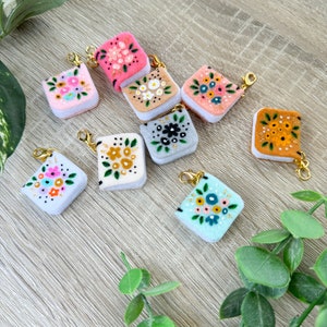 May include: A collection of nine felt mini books with floral embroidery. Each book has a different color and floral design. The books are attached to gold keychains.