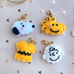May include: Four felt keychains featuring characters from the Peanuts comic strip. The keychains are yellow, white, and black. The characters include Snoopy, Charlie Brown, Woodstock, and Lucy.