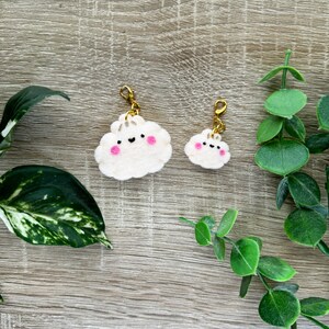 May include: Two white felt cloud keychains with gold clasps. The larger cloud has a pink smiling face with two pink cheeks. The smaller cloud has a pink smiling face. Both clouds are on a light brown wooden background.