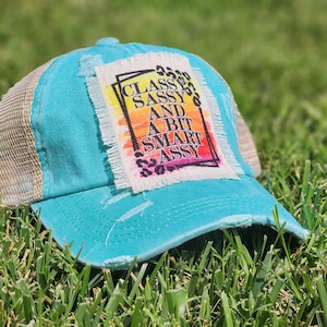 May include: A distressed turquoise trucker hat with a tan mesh back. The front features a rectangular patch with the words "Classy, Sassy, And A Bit Smart Assy" in a colorful design with leopard print accents. The hat is resting on green grass.