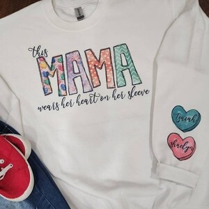 May include: White long-sleeved sweatshirt with the text "This Mama wears her heart on her sleeve" and three colorful heart-shaped patches with the names "Izayah", "Shirley", and "Brian" on them.