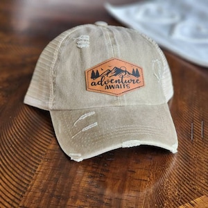 May include: A beige and brown distressed trucker hat with a brown leather patch that says "Adventure Awaits" with a mountain and trees graphic.