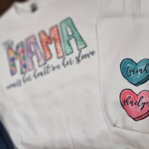 May include: White sweatshirt with the word "Mama" in colorful letters and the text "with her heart on her sleeve". Two heart-shaped patches with the names "Isaiah" and "Shaelyn" are on the sleeve.