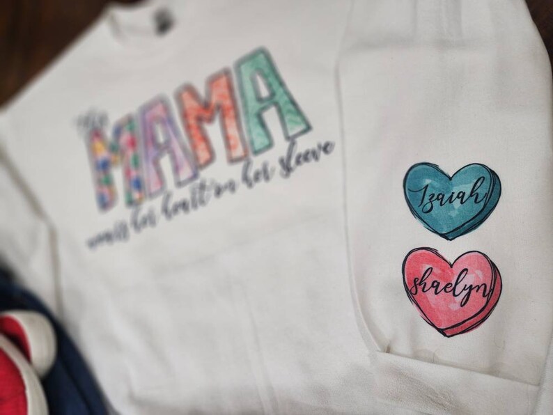 May include: White sweatshirt with the word "Mama" in colorful letters and the text "with her heart on her sleeve". Two heart-shaped patches with the names "Isaiah" and "Shaelyn" are on the sleeve.
