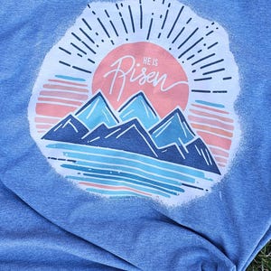 May include: A blue t-shirt with a white graphic of a sunrise over mountains. The graphic includes the text "He is Risen".