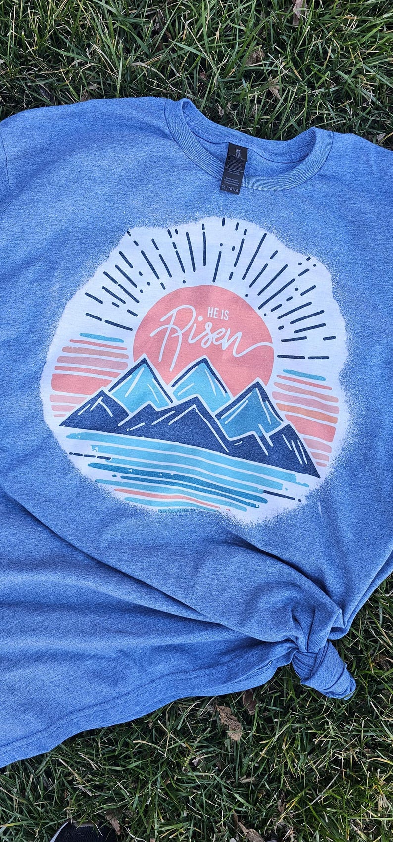 May include: A blue t-shirt with a white graphic of a sunrise over mountains. The graphic includes the text "He is Risen".