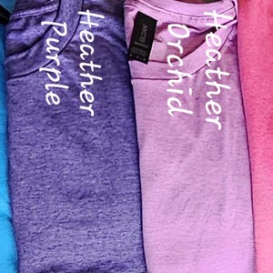 May include: A row of ten t-shirts in different colors, including indigo, teal, sapphire, purple, orchid, pink, and orange. Each shirt has the word "Heather" printed on it, followed by the color name.