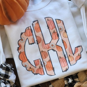 May include: White sweatshirt with a monogram design featuring the letters "CJL" in a circle. The monogram is filled with a pattern of orange pumpkins and white flowers.