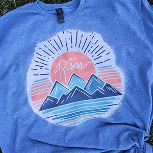 May include: A blue t-shirt with a white distressed graphic of a sunrise over mountains. The graphic includes the text "He is Risen".