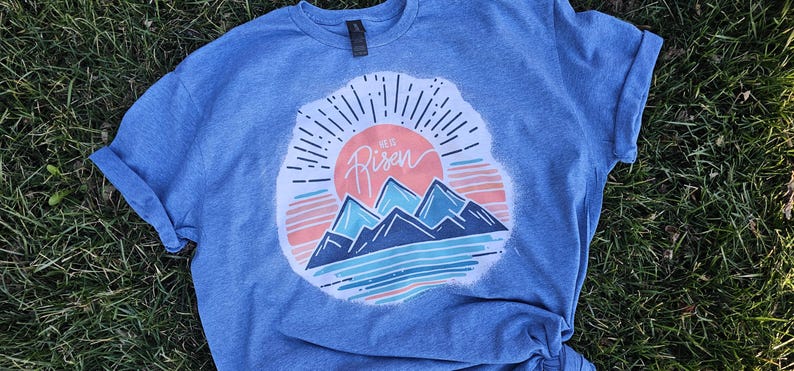 May include: A blue t-shirt with a white distressed graphic of a sunrise over mountains. The graphic includes the text "He is Risen".