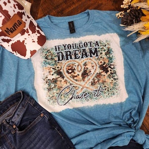 May include: A teal blue t-shirt with a white graphic print that says "If you got a dream chase it". The graphic features a heart shape made of rope and a splatter paint background.
