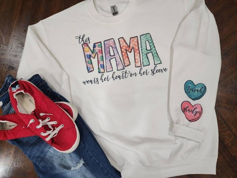 May include: White long-sleeved sweatshirt with the text "This Mama wears her heart on her sleeve" and three colorful heart-shaped patches with the names "Izayah", "Shirley", and "Brian" on them.