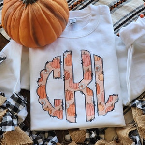 May include: White sweatshirt with a monogram design featuring the letters "C K L" surrounded by a pattern of orange pumpkins and white flowers.