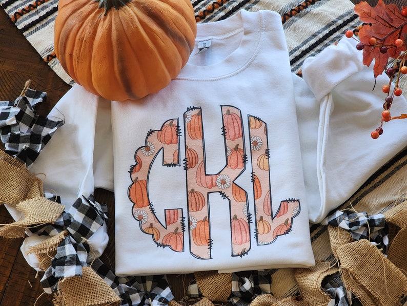 May include: White sweatshirt with a monogram design featuring the letters "C K L" surrounded by a pattern of orange pumpkins and white flowers.