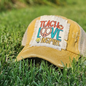 May include: A yellow distressed trucker hat with a white patch that says "Teach Love Inspire" in colorful lettering. The patch has a peace sign, a lightbulb, and an apple.