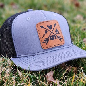 May include: A gray and black trucker hat with a brown leather patch featuring a Mickey Mouse silhouette, a campfire, a camper, and arrows. The patch is stitched onto the front of the hat.