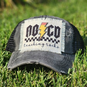 May include: Black distressed trucker hat with a white patch that says "ABC" with a yellow lightning bolt and "teaching rocks" in black script.