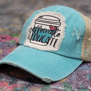 May include: A turquoise blue and beige trucker hat with a distressed look. The hat has a white patch with a coffee cup graphic and the text "caffeinate & educate".