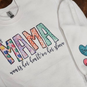 May include: White sweatshirt with the text "This mama wears her heart on her sleeve" and colorful patterned letters spelling "mama". The sleeves have two heart-shaped patches with the names "Isaiah" and "Shaelyn" written on them.