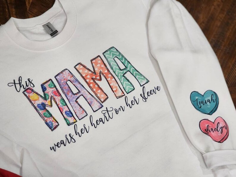 May include: White sweatshirt with the text "This mama wears her heart on her sleeve" and colorful patterned letters spelling "mama". The sleeves have two heart-shaped patches with the names "Isaiah" and "Shaelyn" written on them.