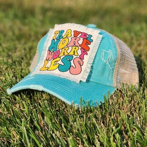 May include: A turquoise blue trucker hat with a white patch that says "Travel More Worry Less" in colorful, retro-style lettering.