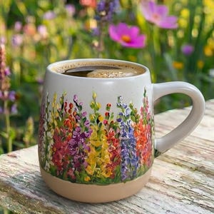 May include: A ceramic mug with a speckled off-white finish featuring a hand-painted design of colorful wildflowers in shades of purple, yellow, red, and blue around the base. The mug has a curved handle and contains a dark brown liquid beverage.