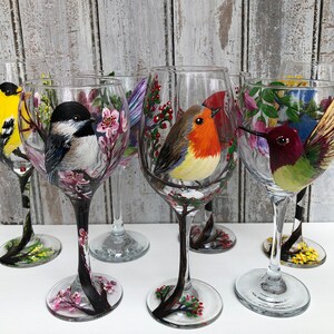 May include: Six clear wine glasses, each hand-painted with a different colorful bird design. The birds include a yellow finch, a black-capped chickadee, a robin, and a hummingbird. The stems are painted black with floral accents.