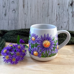 May include: A white ceramic mug with a handle, adorned with hand-painted purple flowers with yellow centers and green leaves. A small bouquet of purple flowers sits beside the mug on a wooden surface.