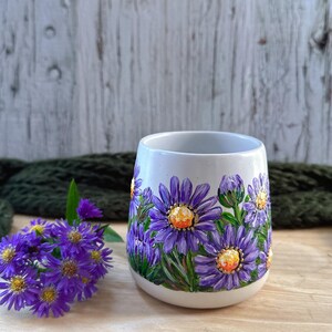 May include: A white ceramic mug with hand-painted purple aster flowers and green leaves. The flowers have yellow centers. A small bouquet of purple flowers sits to the left of the mug. The mug is on a wooden surface.