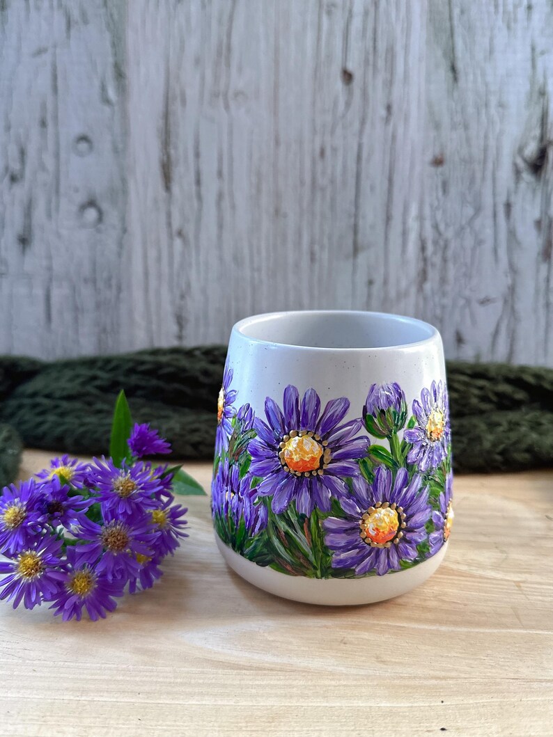 May include: A white ceramic mug with hand-painted purple aster flowers and green leaves. The flowers have yellow centers. A small bouquet of purple flowers sits to the left of the mug. The mug is on a wooden surface.