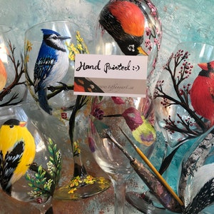May include: Hand-painted wine glasses featuring detailed bird illustrations. The glasses showcase a variety of colorful birds, including a cardinal, blue jay, and goldfinch, perched on branches with floral accents. The text "Hand Painted" is visible.