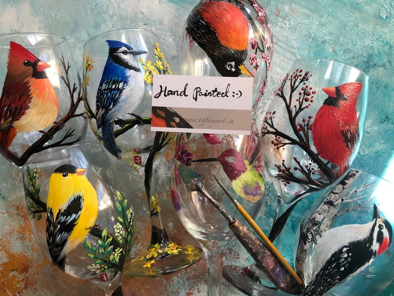 May include: Hand-painted wine glasses featuring detailed bird illustrations. The glasses showcase a variety of colorful birds, including a cardinal, blue jay, and goldfinch, perched on branches with floral accents. The text "Hand Painted" is visible.
