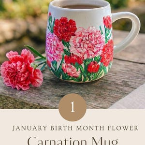 May include: A ceramic mug featuring hand-painted red and pink carnation flowers with green stems and leaves. A single fresh pink carnation rests beside the mug. The text reads: 1 JANUARY BIRTH MONTH FLOWER Carnation Mug HAND PAINTED WITH LOVE.