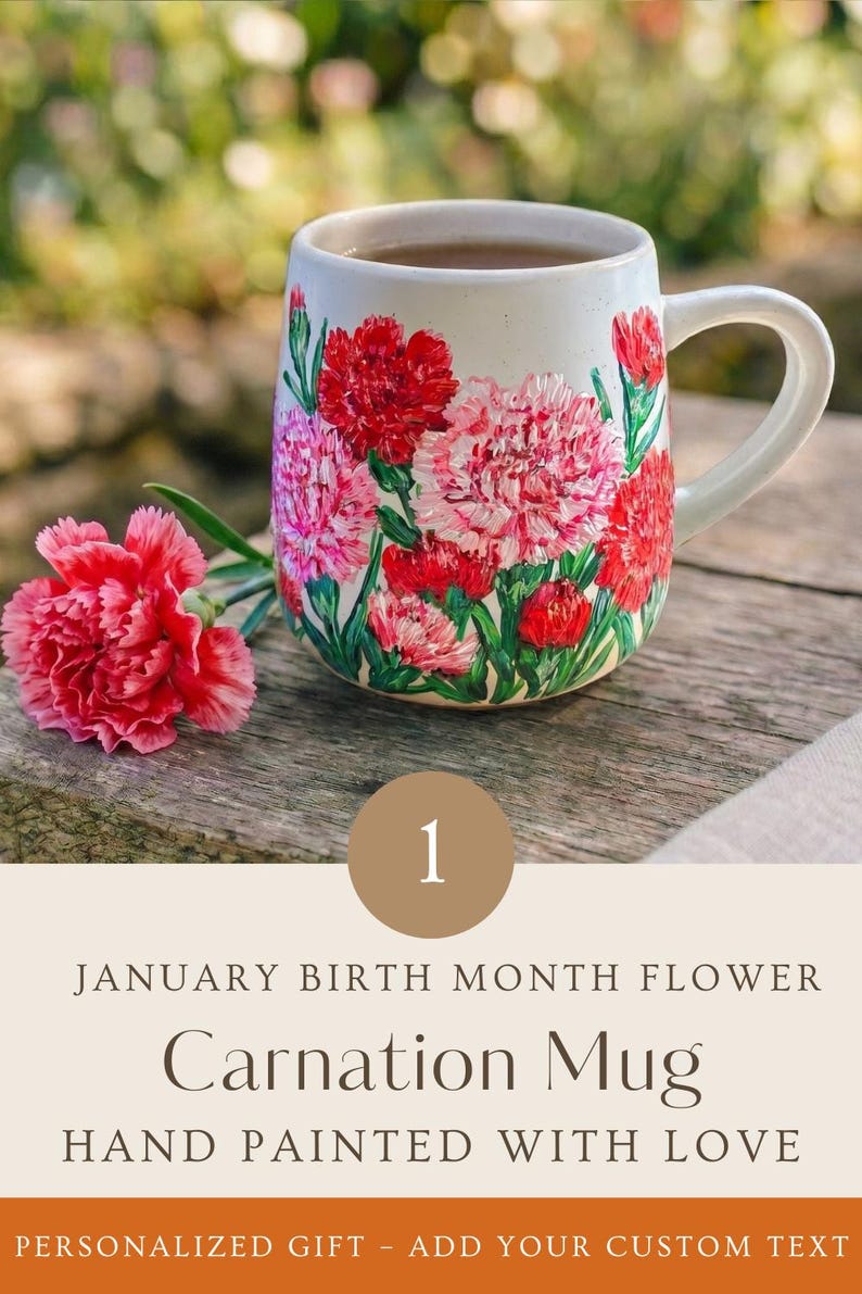 May include: A ceramic mug featuring hand-painted red and pink carnation flowers with green stems and leaves. A single fresh pink carnation rests beside the mug. The text reads: 1 JANUARY BIRTH MONTH FLOWER Carnation Mug HAND PAINTED WITH LOVE.