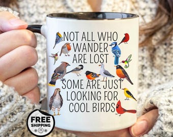 May include: White ceramic mug with a black rim and handle, featuring colorful illustrations of various birds. The mug has the text "NOT ALL WHO WANDER ARE LOST SOME ARE JUST LOOKING FOR COOL BIRDS".