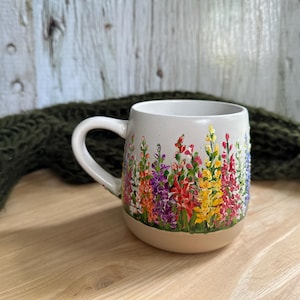 May include: A ceramic mug with a speckled off-white finish featuring a hand-painted design of colorful lupine flowers in shades of red, purple, yellow, and pink. The mug has a rounded body, a curved handle, and a smooth, unglazed tan base.