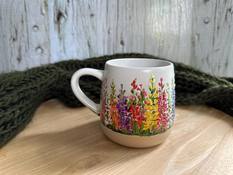May include: A ceramic mug with a speckled off-white finish featuring a hand-painted design of colorful lupine flowers in shades of red, purple, yellow, and pink. The mug has a rounded body, a curved handle, and a smooth, unglazed tan base.
