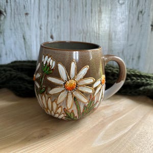 Pode incluir: Uma caneca de cerâmica marrom com um design floral de margaridas brancas. As margaridas têm centros laranja e caules verdes. A caneca tem uma forma arredondada e uma alça.
