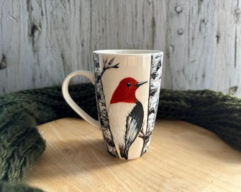 May include: White ceramic mug with a hand-painted design of a red-headed woodpecker perched on a birch tree branch. The bird has black and white markings on its wings and tail.