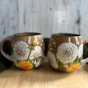 Può includere: Due tazze in ceramica marrone con disegni di dente di leone bianchi e gialli. Le tazze hanno una forma arrotondata e un manico.